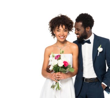 Smiling bride holding flower bouquet while groom looks at her with a smile